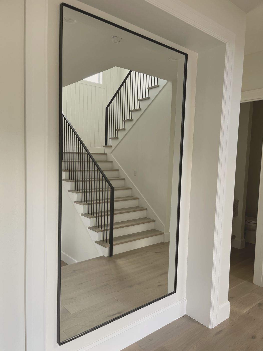 A black FRAMED steel angle wall piece in a white hallway, reflecting a staircase and its steel railing.